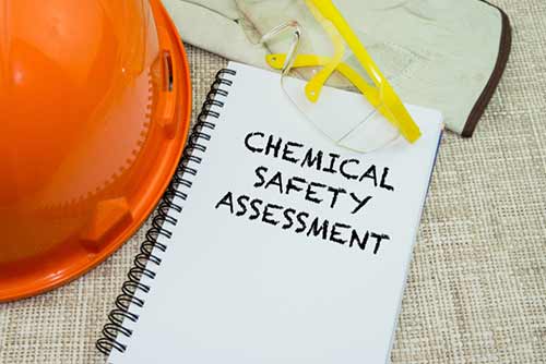 safety equipment inclduing a hard hat glasses and gloves laying next to a pad that reads chemical safety assessment
