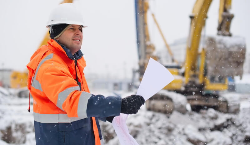 worker in high visibility workwear surveying in the snow next to a large digger