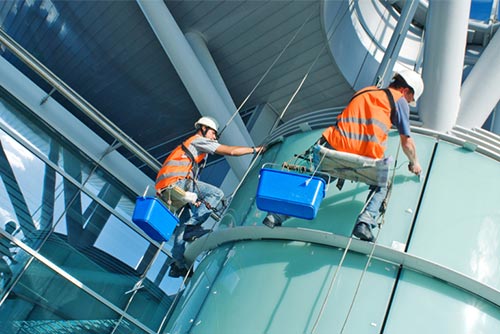 two men cleaning windows on a high rise building suspended with cables wearing orange high vis vests